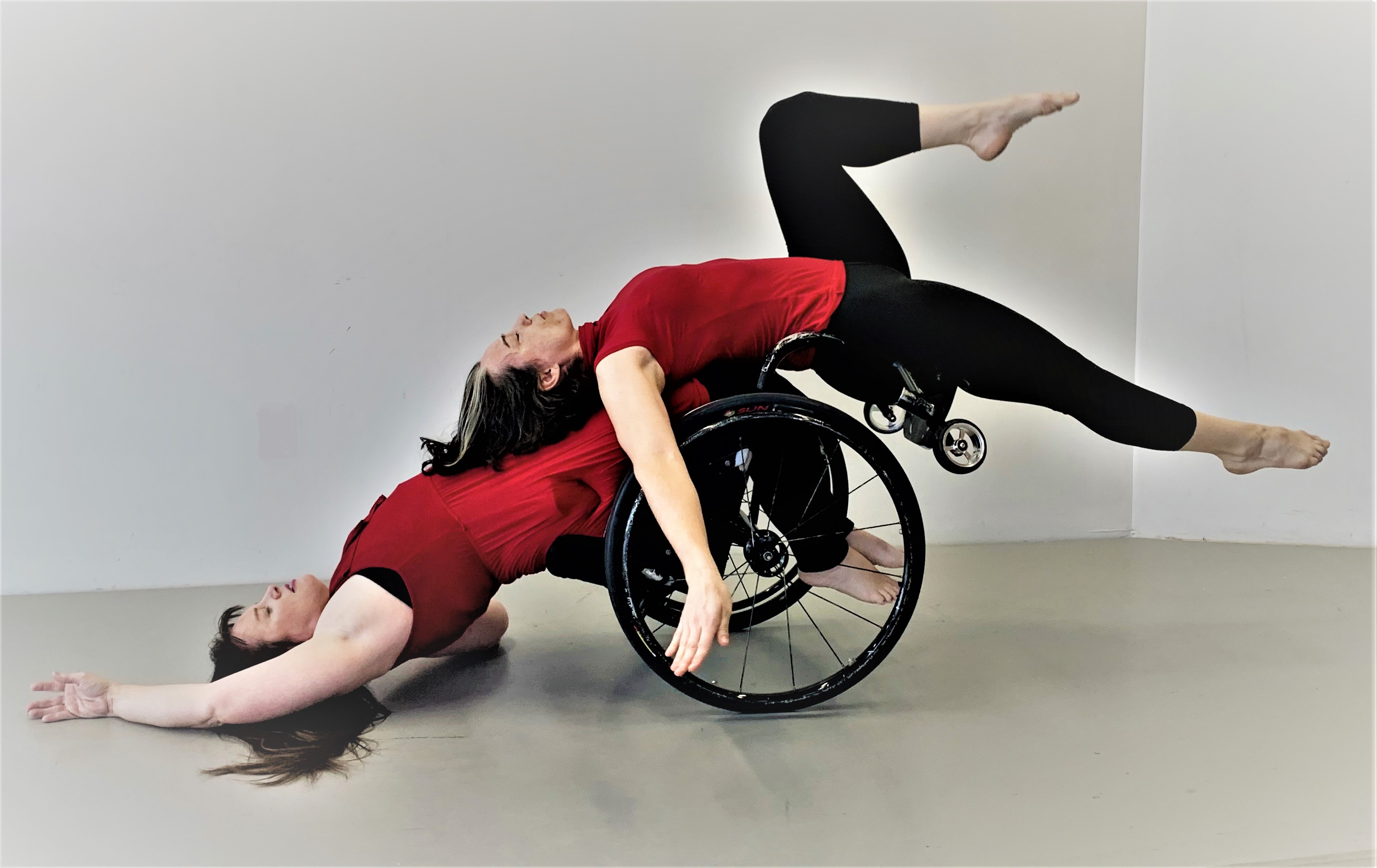 Lindy, a white woman with long brown hair, arches back in her wheelchair with her shoulders barely grazing the ground. Ashlee, a white woman with dark hair, counterbalances Lindy by laying on her footplate and knees. Both are parallel to the floor, creating a pose that suggests Ashlee is using her body weight to return Lindy to an upright position in her chair. The dancers wear red tops and black leggings.