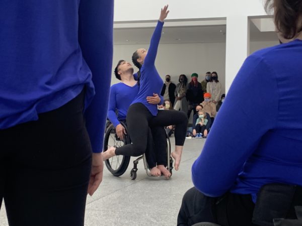 Pictured: In the atrium of the High Museum of Art is Peter, a mixed race male half Asian, half Caucasian in his 30s with medium length dark hair, scruffy beard, using a manual chair. On his lap sits Ashlee, a tall, strong-bodied white woman. She has dark hair, brown eyes and freckles. Ashlee is reaching her right arm towards the sky. They are clad in navy blue long-sleeved shirts and black pants.