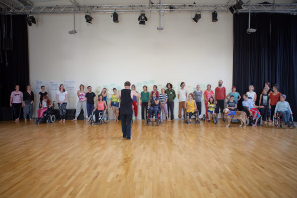 Douglas, an older nondisabled white man, stands with his back to the camera, wearing a black sleeveless shirt and black pants. In a line that spans the photo from left to right, dancers of various embodiments are facing Douglas, listening to his instructions – including one canine assistant.