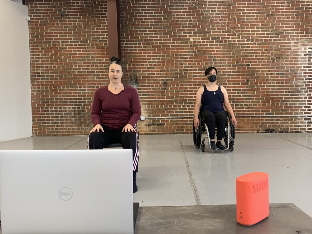 Ashlee, a tall white womand with dark hair, is seated in a folding chair. Beside her sits Peter, a disabled mixed-race male in a manual wheelchair. The background is the red brick wall of the dance studio.