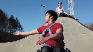 Peter, a mixed race male in his 30s, sits in his wheelchair with his arms gesturing to the side. In the background, Juliiana, a white woman in her 20's, sits high above Peter on a concrete ramp. 