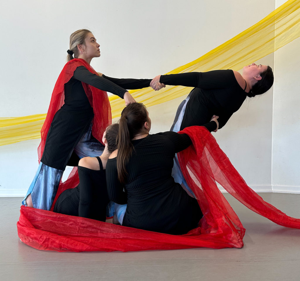 Four dancers, two standing, two seated one the floor, create a geometric trapezoid shape