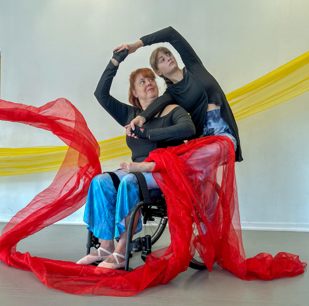 Two dancers, one standing and one in a manual wheelchair, intertwine their arms. The standing dancer is behind the seated dancer.