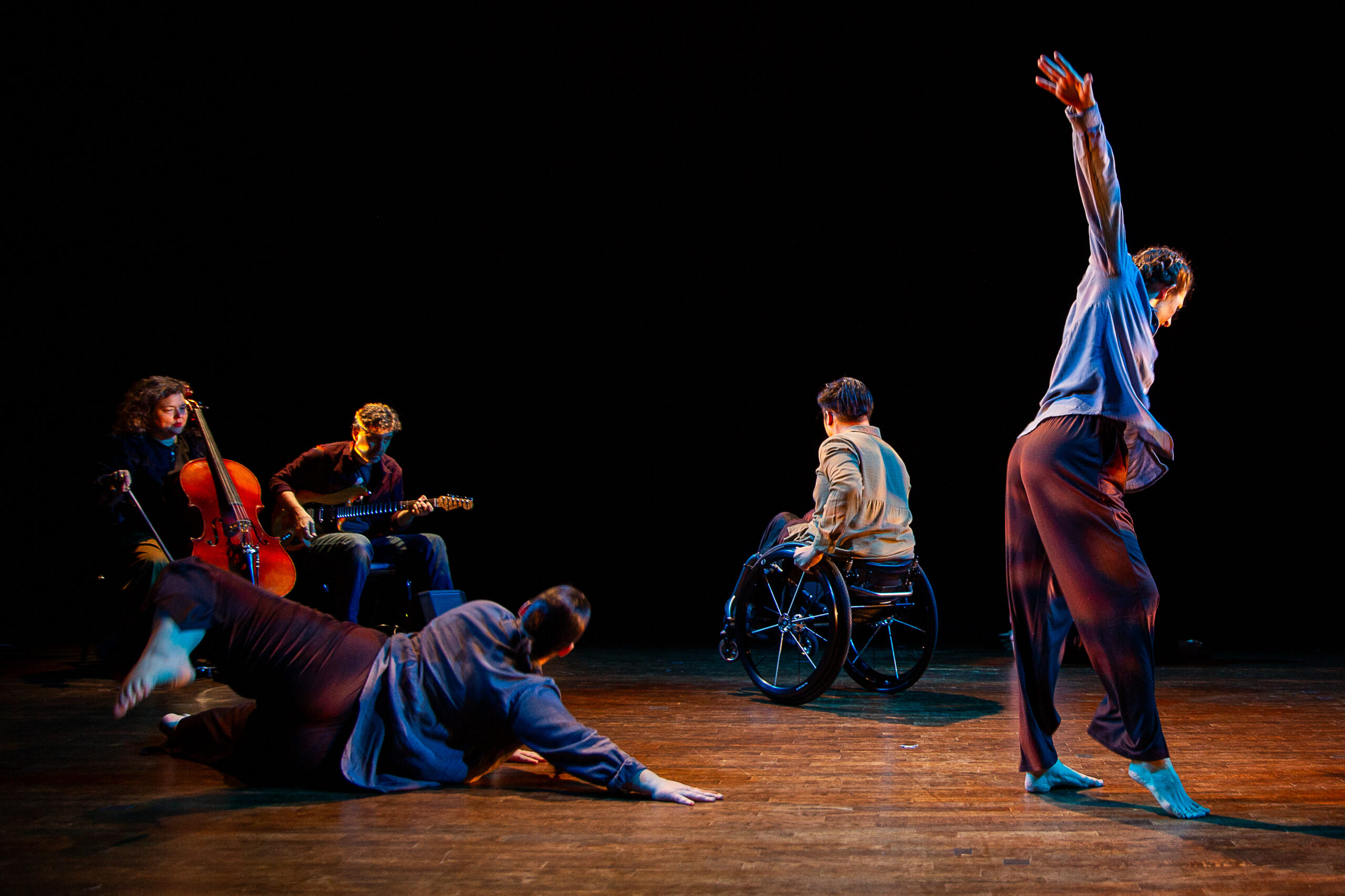 A cellist and a guitarist are picutured on the right of the photo. Three dancers, one on the floor, one in a manual wheelchair, and one standing are caught mid motion.