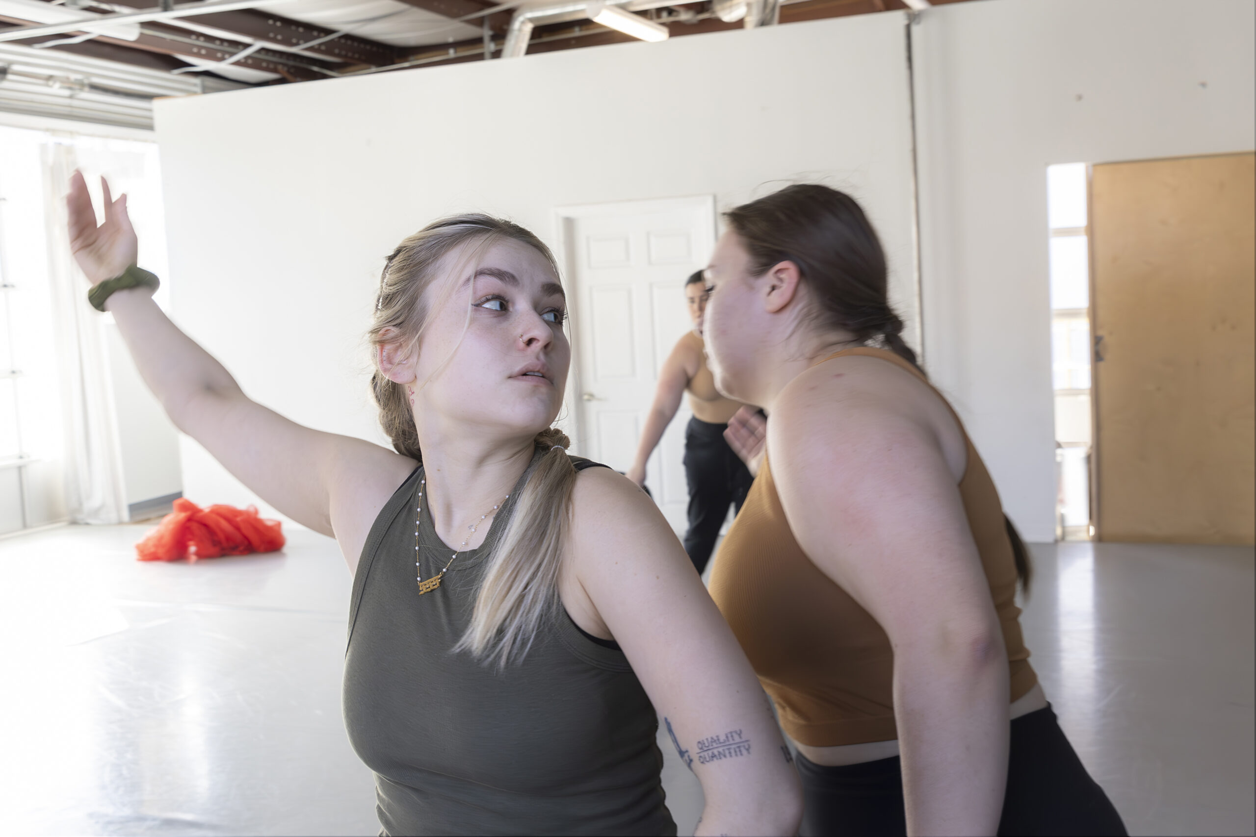 AK Bayer, a nonbinary, disabled dancer looks to the left. They are lifting their right arm to the side. Behind AK, Cecilia, a tan skinned Hispanic disabled woman, leans away from AK.