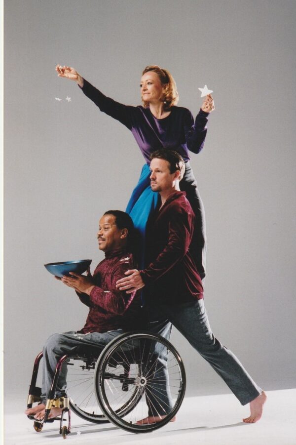 Ardath, a white woman with blonde hair, sits on Douglas' shoulder. She holds a star in her extended right arm. Douglas, a white man with a goatee, rests his hands on Tommy's shoulders. Tommy, a black man with a mustache, sits in his manual wheelchair holding a wooden bowl full of stars.