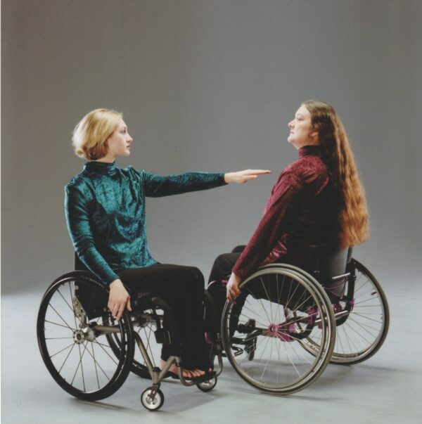 Teal Sherer, a white woman with short blonde hair who uses a wheelchair, extends her arm towards Jaehn Clare, almost touching her. Jaehn is a white woman with long brown hair that extends down her back. Jaehn is seated in her manual wheelchair.