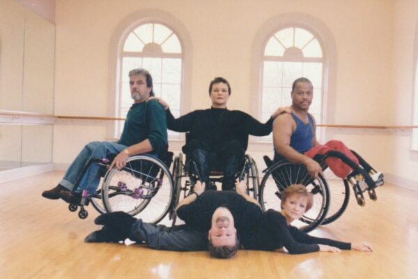 Gary, an white male with a mustache and beard, is in wheelie with his knees facing to the right. Sandra, a mixed race female, is facing front with her hand on his shoulder. Her other hand rests on Tommy, a black male in a wheelie with his knees facing to the left. On the floor in front of this trio are Douglas, a white male with a goatee, and Ardath, a white female with blonde hair. Douglas lies on his back, his legs under Sandra's wheelchair. He holds her in a wheelier. He arches his back over Ardath, who lies on the floor.
