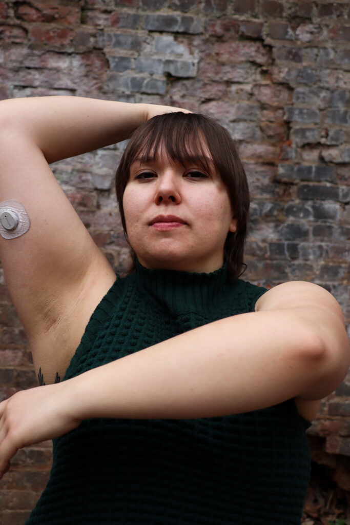 Cecilia is a white Latina woman with brown hair and brown eyes. In the photo, Cecilia wears a dark green turtleneck tank top. Her hair is down and her bangs in front of her face. She poses with one arm bent above her head and the other curved around the front of her body. On her arm, you can see her Dexcom continuous glucose monitor. Not pictured is her insulin pump.