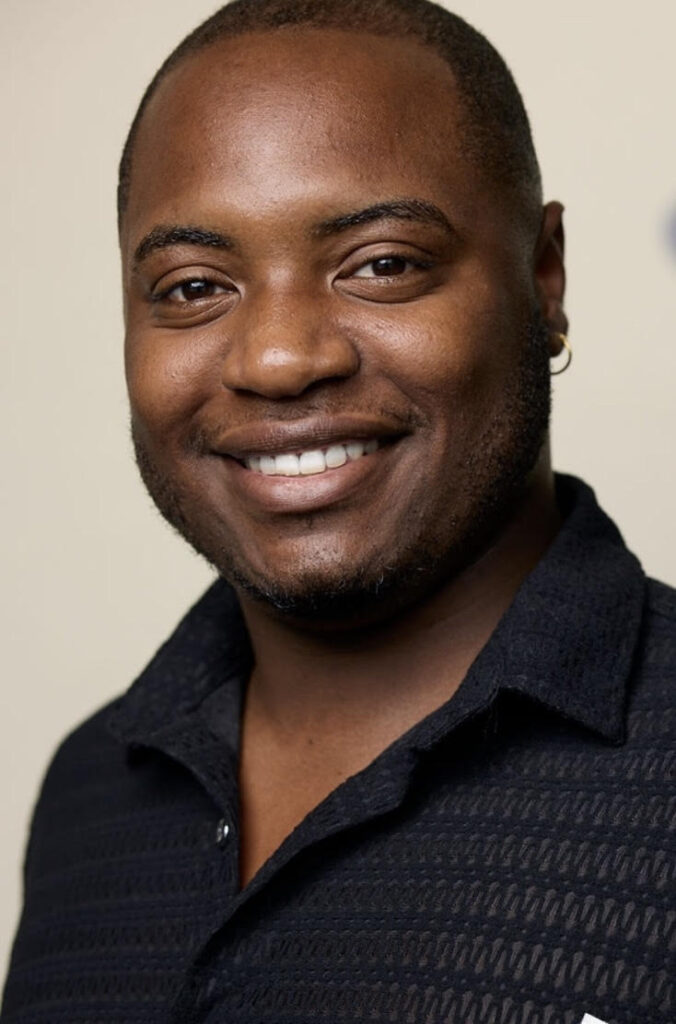 Delaronce is a African American malewith a low haircut and a beard. Delaronce is wearing a black chevron button down  and has an enthusiastic smile.