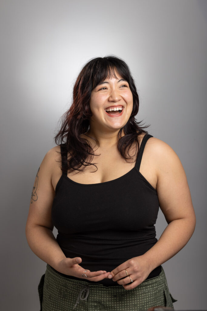 Hiroko is a biracial Asian and White female identifying woman with black hair, brown eyes, and light freckles across her face. In this photo, she has medium length hair with bangs, wearing a black tank top, and is looking off to the side smiling joyfully.