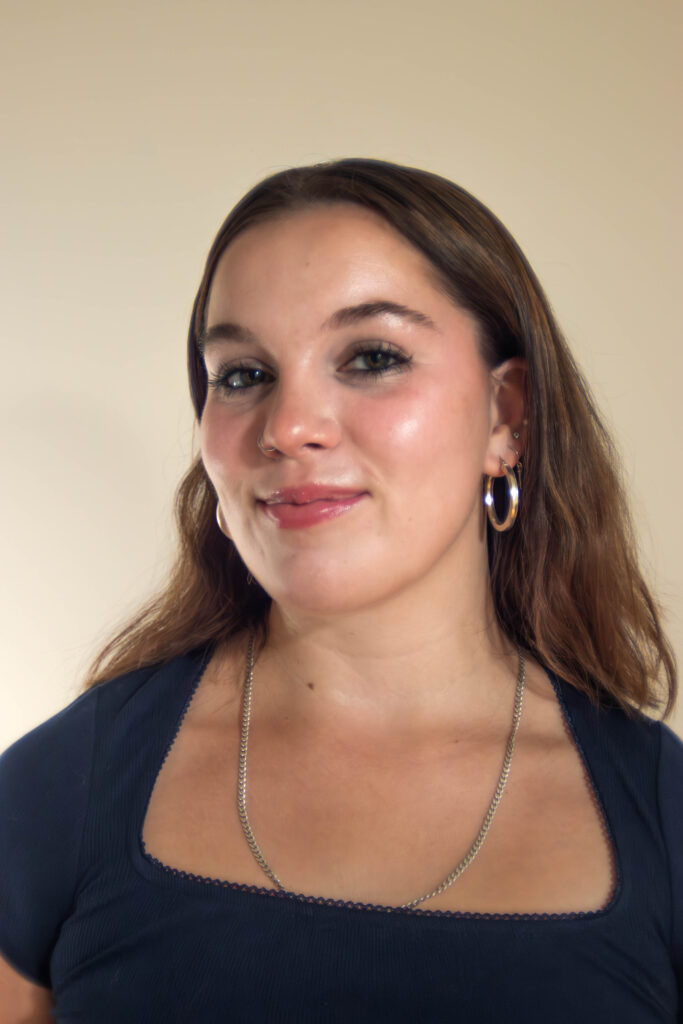 Madeline is a white woman with brown hair. She wears hoops and a silver necklace. She smiles softly with her head tilted to the right. 