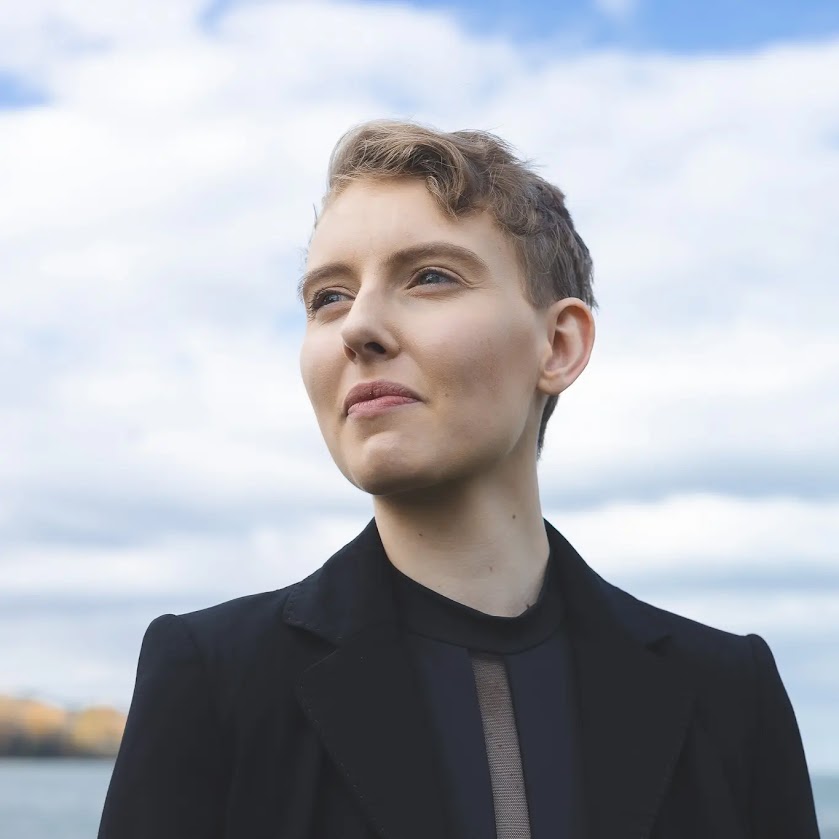 Trinity is a white person with short blonde hair. They are wearing a black suit jacket with a big smile on their face. They look slightly to the side against a backdrop of blue sky.