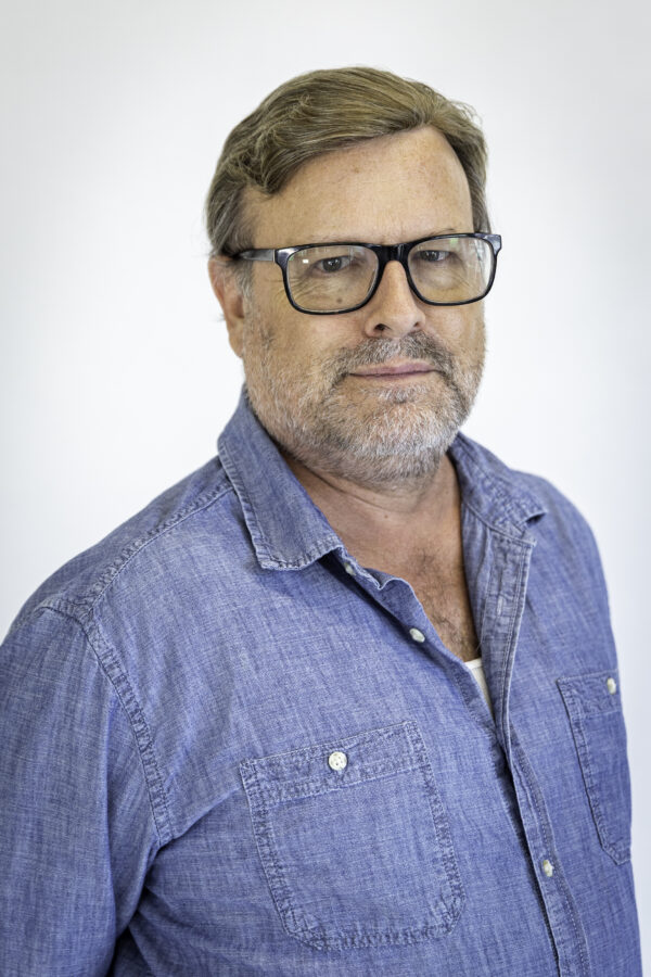 Douglas is an older white male with a closely cropped grey beard, glasses, and brown hair. He is wearinga blue shirt.